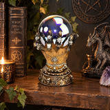 A decorative crystal ball on a silver and gold gothic hand sculpture stand, displayed on a wooden shelf next to a lit candle, old spellbook, and a dragon figurine.