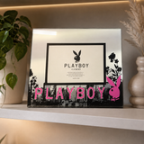 A rectangular framed mirror or picture frame displayed on a shelf, featuring a black Playboy bunny logo and the word PLAYBOY at the bottom, with a pink Playboy bunny head accent and floral details. The frame is shown in a styled interior setting with a plant and vase on a shelf.