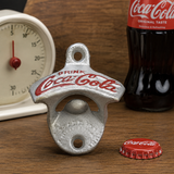 A silver metal bottle opener shaped like a Coca-Cola bottle cap, with red Coca-Cola branding, standing on a wooden surface. A red Coca-Cola bottle and a white clock are blurred in the background.